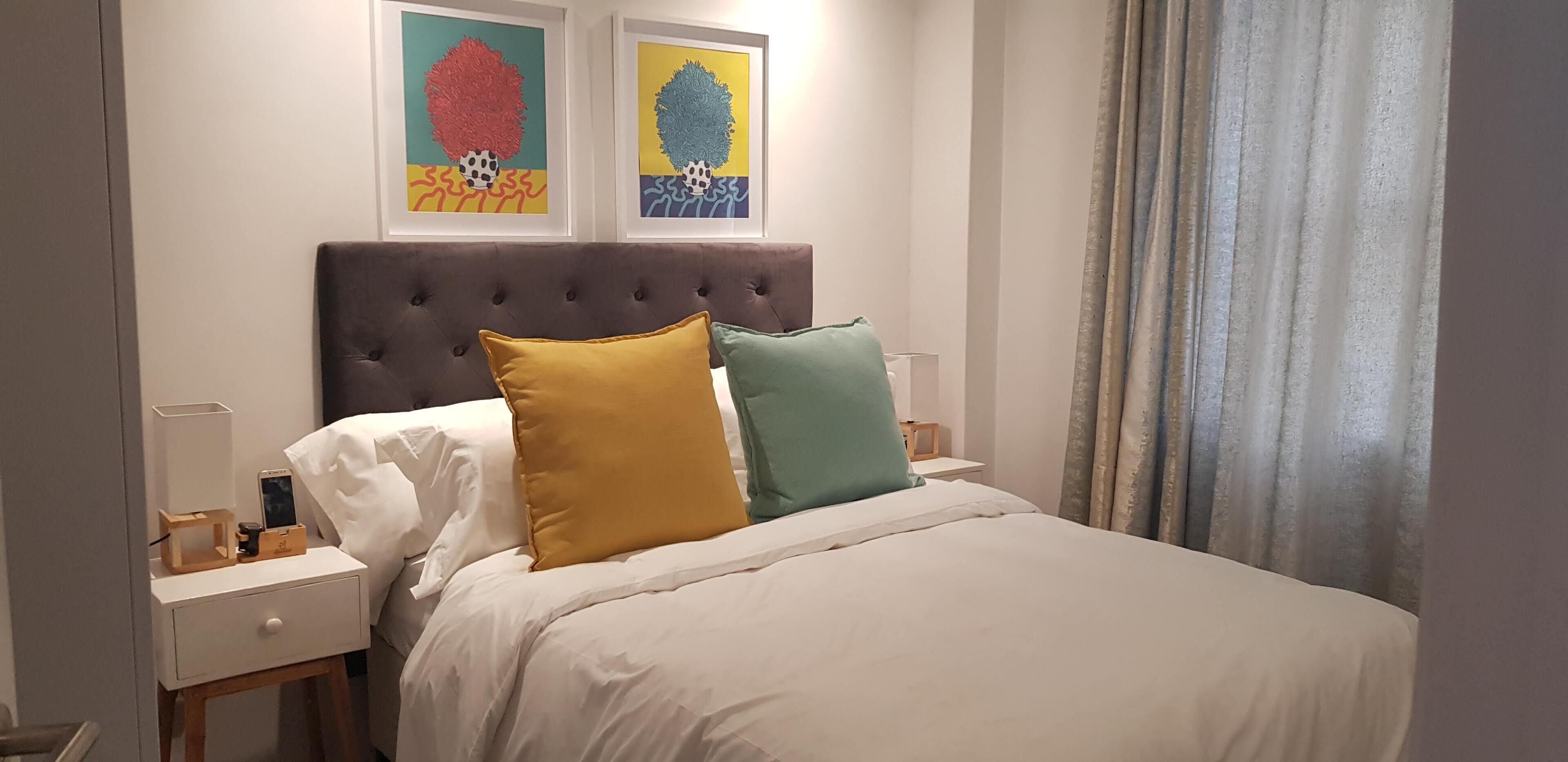 Een netjes opgemaakt bed met witte lakens, twee kussens in mosterdgeel en blauwgroen, een houten nachtkastje met een lamp, en twee ingelijste kunstwerken erboven.