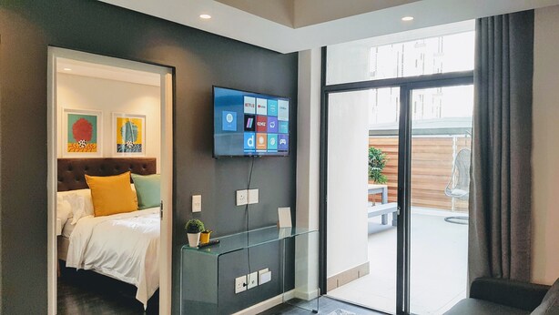 Ein modernes Hotelzimmer mit einem Bett, einem an der Wand befestigten Fernseher, der Streaming-Dienste anzeigt, einer Glasvitrine und einer Schiebetür zu einem Außenbereich.