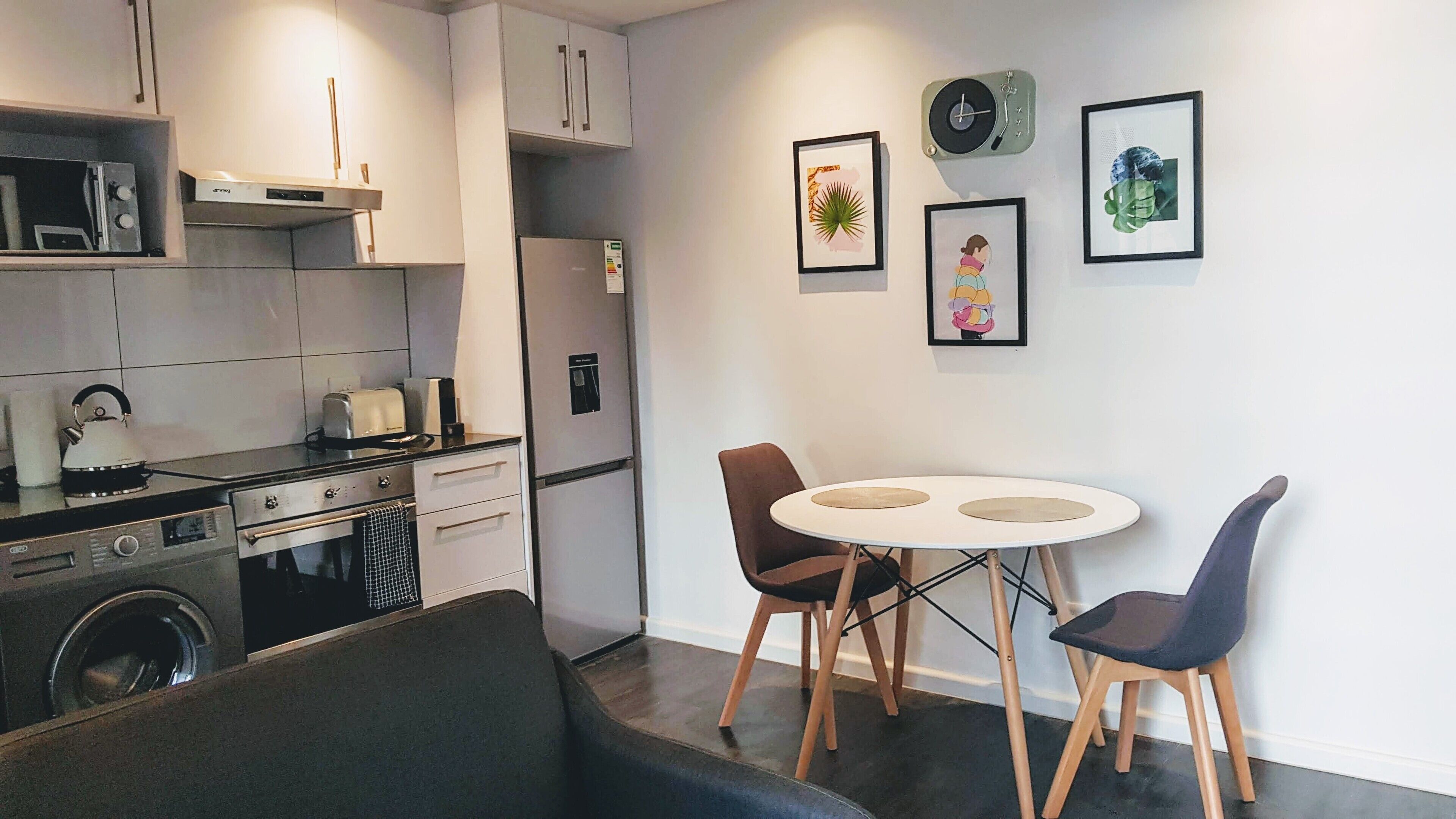A compact kitchen with modern appliances, a dining area with two chairs and a small table, and a wall with framed artwork.