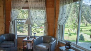 A cozy room with two leather chairs, a small table with a lamp, and a view of a green landscape through large windows. - Eagle's Nest For Great Hikes, Local Breweries and Coffee Shops. (Lolo)
