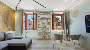 A modern living room with a sectional sofa, two chairs, a stone wall with a painting, and a view of a building through open windows. - Diana City Hall View (Pula)