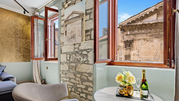 A room with a stone wall, a window with a view of a historic building, a round table with a bottle and flowers, and a chair. - Diana City Hall View (Pula)