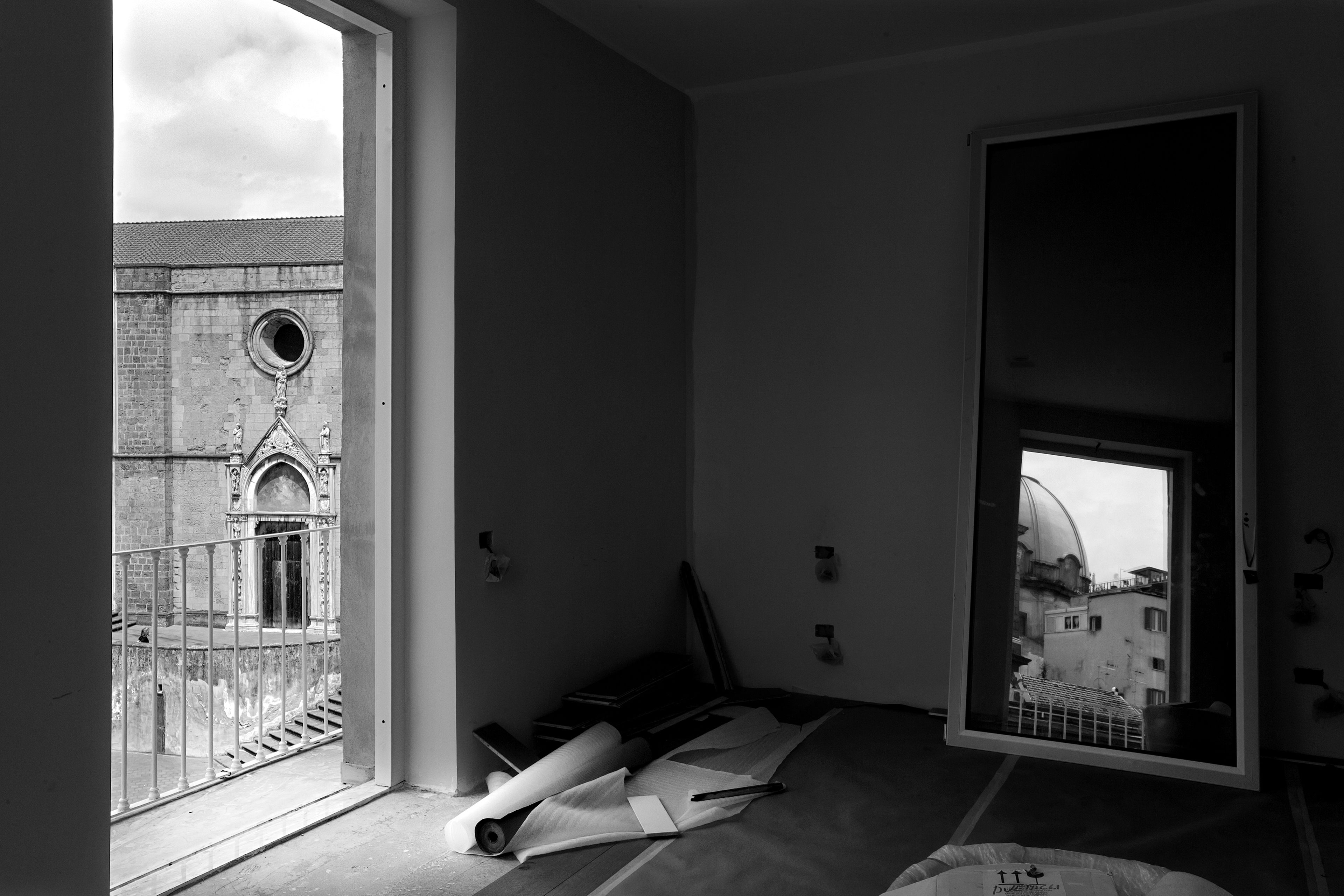 A room with a large window overlooking a historic building with a circular window and arched doorway. The room has a carpeted floor and a bed with a white headboard.