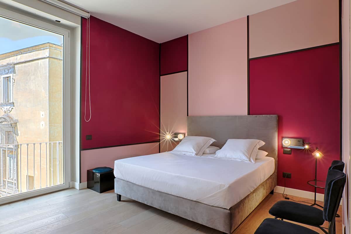 A bedroom with a large bed, a nightstand, a chair, a desk, and a window with a view of a building.