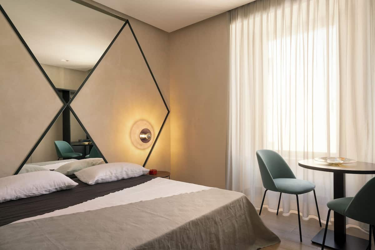 A modern hotel room with a bed, two chairs, a table, and a large mirror.