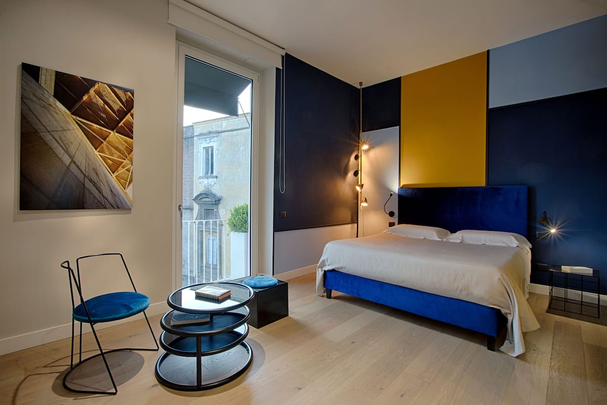 A modern bedroom with a large bed, a blue chair, a round table, and a city view through a large window.