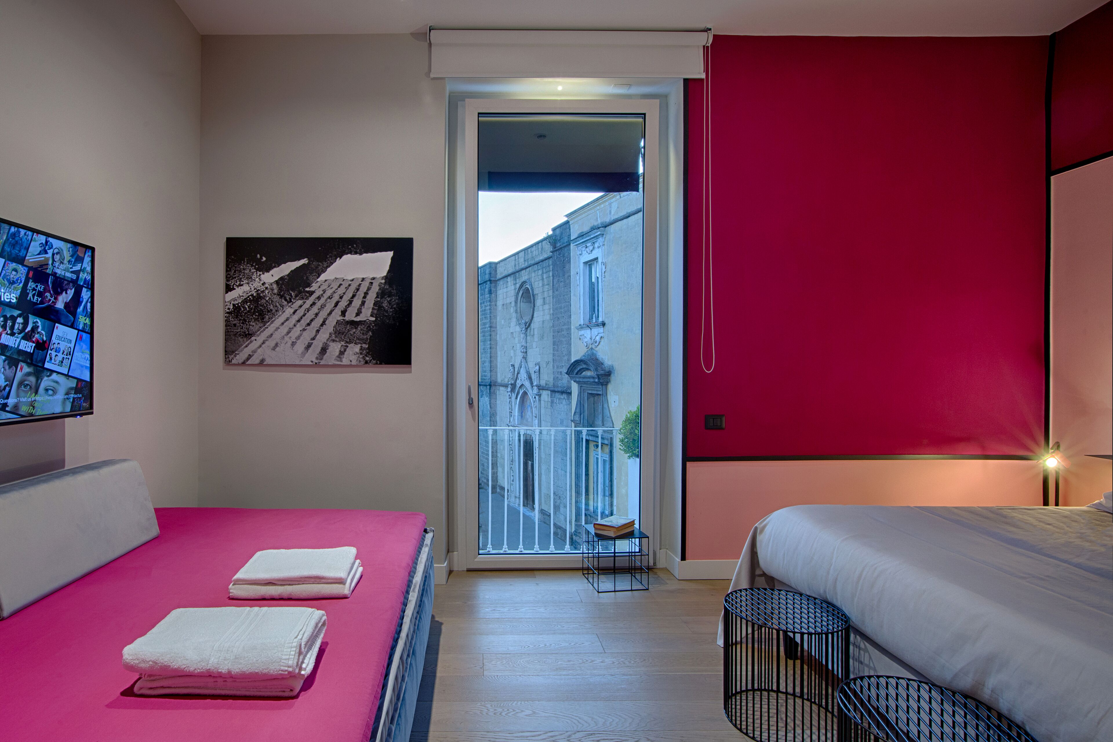 A hotel room with a bed, a TV, a painting, and a view of a building through a glass door.