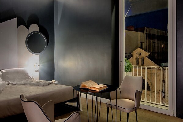 A modern bedroom with a bed, a round mirror, a small table with an open book, and a chair. A balcony with a railing and a view of a building is visible. - Casa pacifico Napoli (Naples)