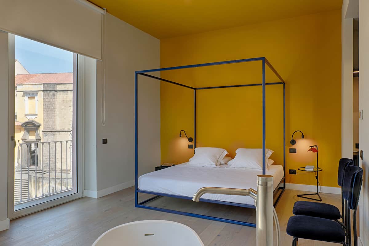 A modern bedroom with a four-poster bed, a large window, a small bedside table, and a chair.