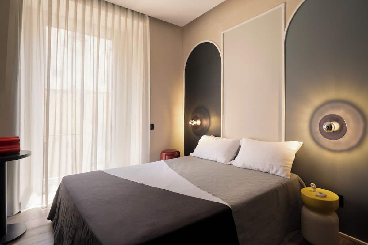A modern hotel room with a large bed, a bedside table, and a small stool.