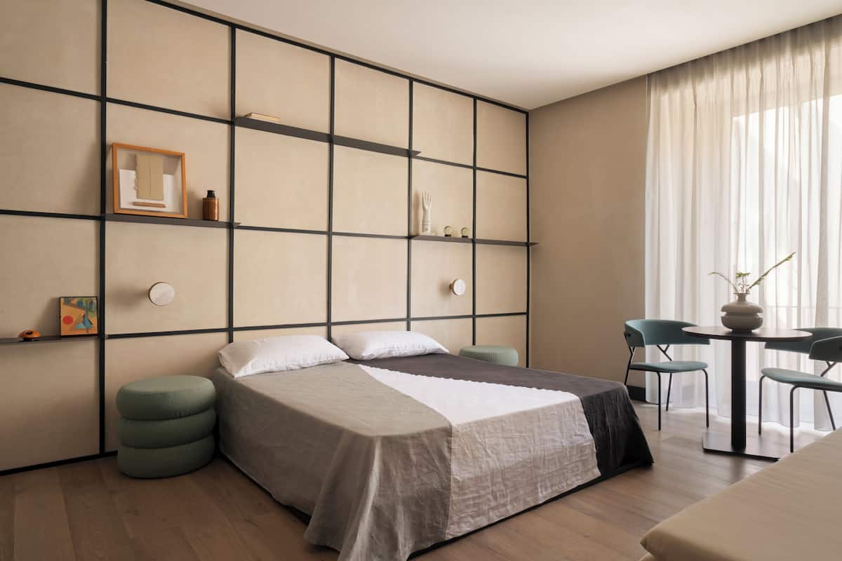 A modern hotel room with a large bed, a small table with chairs, and a view of the outdoors through sheer curtains.