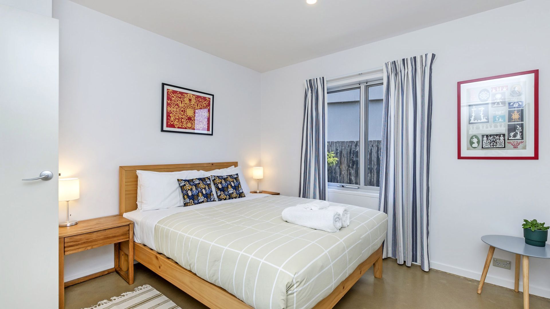 A bedroom with a bed, bedside tables, a window with curtains, a wall-mounted artwork, and a small table with a plant.