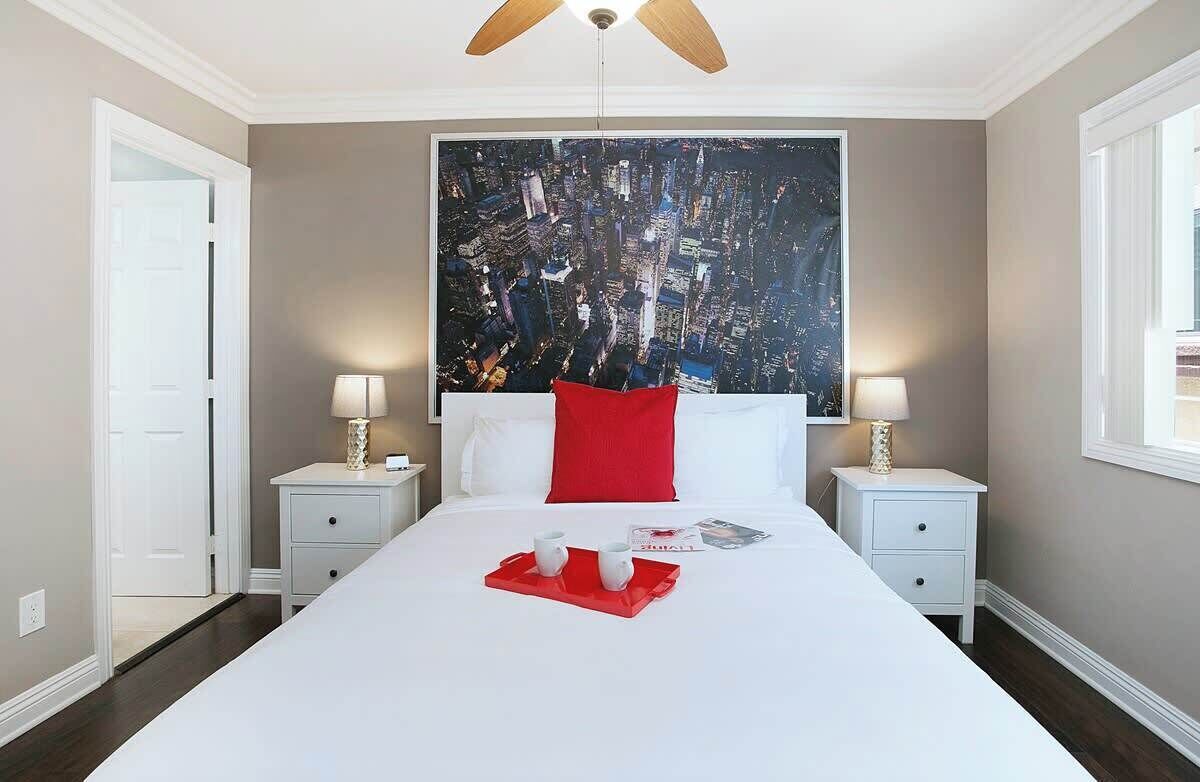 A hotel room with a large bed, two bedside tables with lamps, a red pillow, and a cityscape painting on the wall.
