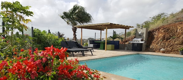 Una piscina con sillones de descanso, zona de barbacoa y vegetación circundante.