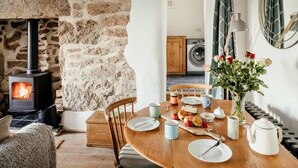 A cozy living room with a stone wall, a fireplace, a dining table set with plates, cups, and a vase of flowers. - 17 Farmers Meadow (Penzance)