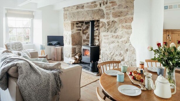 A cozy living room with a stone fireplace, a comfortable sofa, a dining table set for four, and a television. - 17 Farmers Meadow (Penzance)