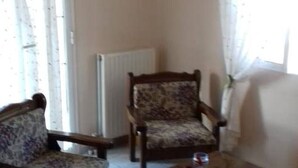 Ein Zimmer mit zwei Sesseln, einem kleinen Tisch, einem Heizkörper und einem Fenster mit Vorhängen.