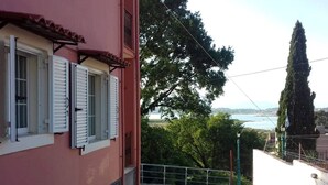 Ein rosafarbenes Gebäude mit weißen Fensterläden, Blick auf ein Gewässer und ein hoher Baum.
