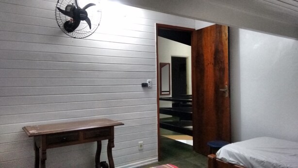 A room with a wooden desk, a ceiling fan, a bed, and a door leading to another room. - Maresias. Spacious and comfortable house. With 4 bedrooms and close to everything. (Sâo Sebastião)