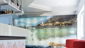 A modern bar area with a glass table, red chairs, and a scenic mural of a coastal landscape. - Hotel Augusta (Malcesine)