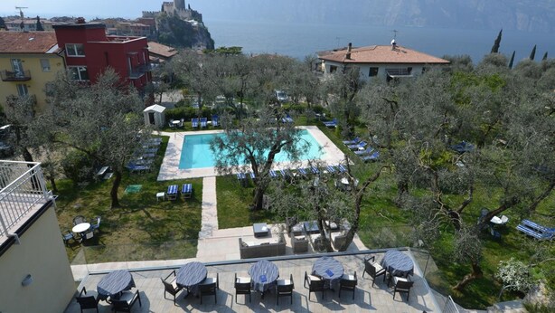 A pool area with lounge chairs and tables, surrounded by trees and buildings, overlooking a body of water and mountains. - Hotel Augusta (Malcesine)