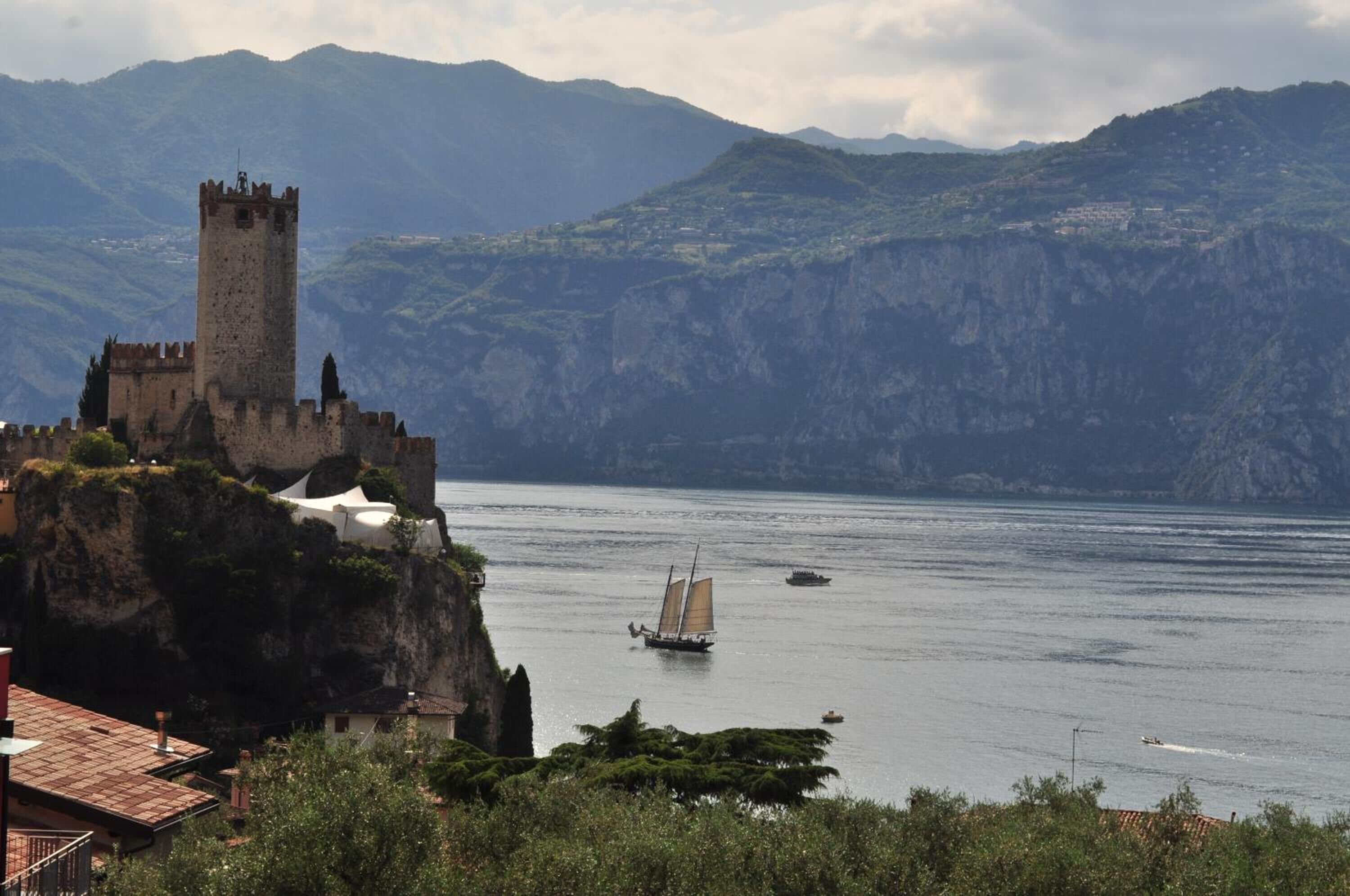 Un castillo costero con una torre, rodeado de montañas y un mar tranquilo con barcos.