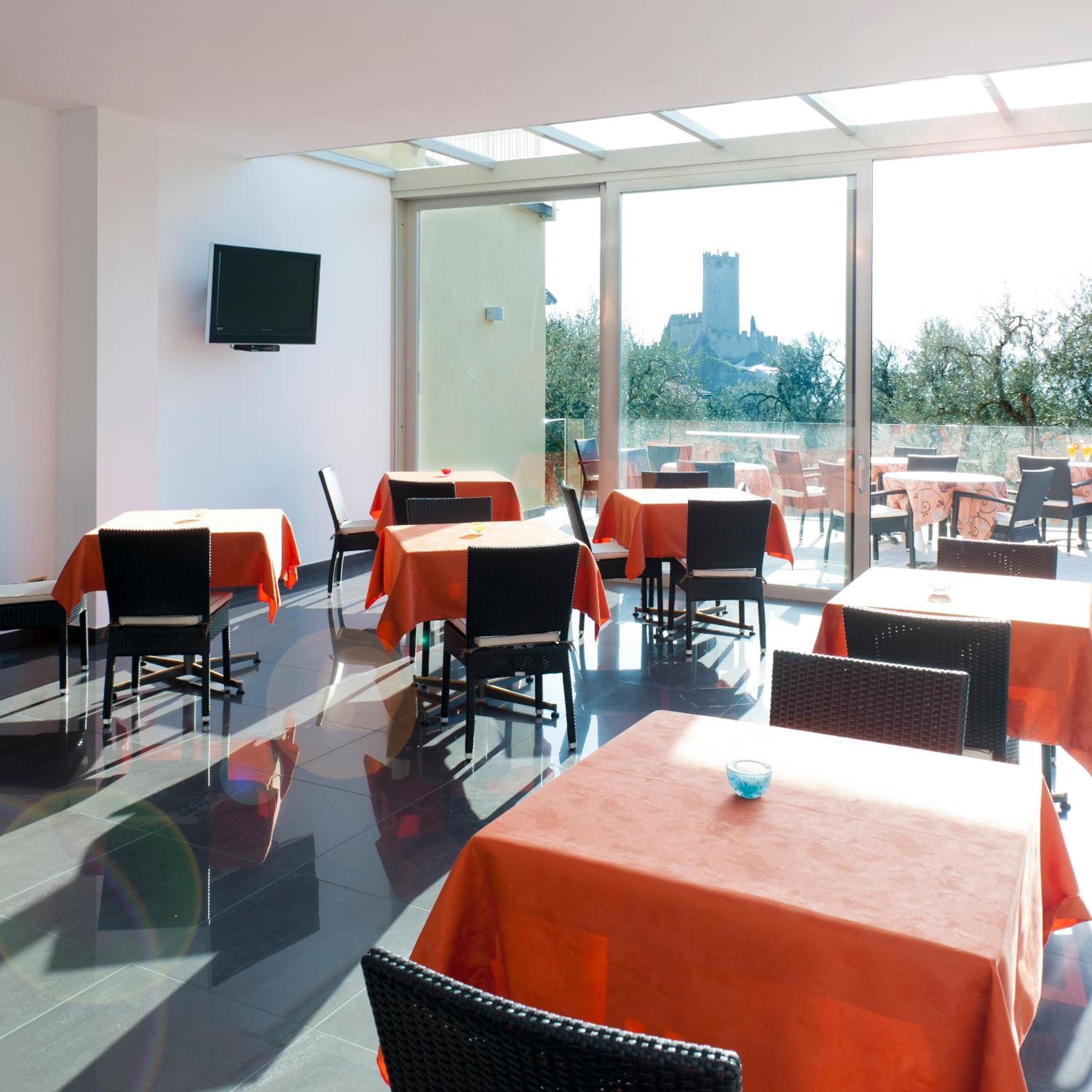 Un restaurant dont les tables sont recouvertes de nappes orange, avec des chaises noires et une grande fenêtre donnant sur un château.