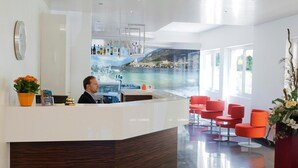 A modern reception area with a counter, a receptionist, and a view of a coastal landscape through a glass wall. - Hotel Augusta (Malcesine)