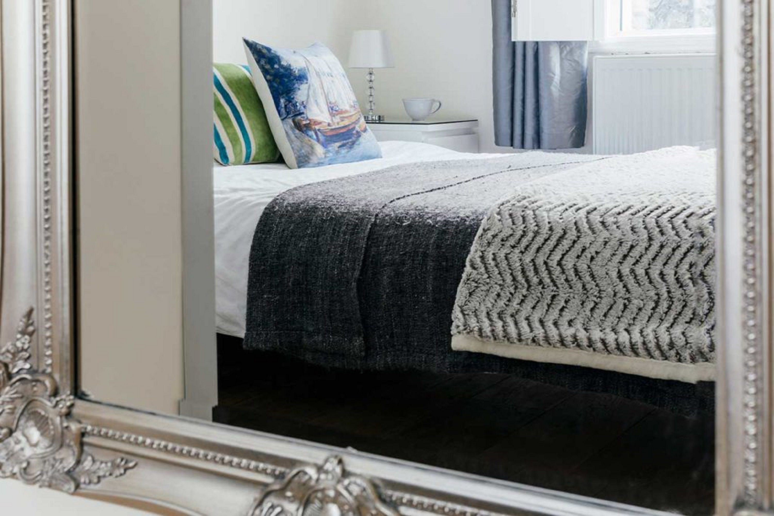 A neatly made bed with a patterned blanket and pillows, a bedside table with a lamp, and a window with curtains.