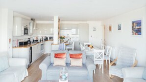 A modern living room with a grey sofa, white armchairs, and a coffee table. There is a kitchen in the background with white cabinets and a dining area with a white table and chairs. - 10 Fernlea Court (St Ives)