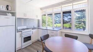 Ett modernt kök med vita skåp, en spis och en diskmaskin. Det finns ett matbord med stolar och ett fönster med utsikt över träd.