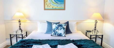 A hotel room with a bed, two bedside lamps, a painting of a beach, and a fluffy green blanket.
