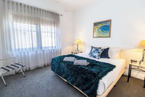A hotel room with a bed, two bedside lamps, a painting on the wall, and a window with sheer curtains.