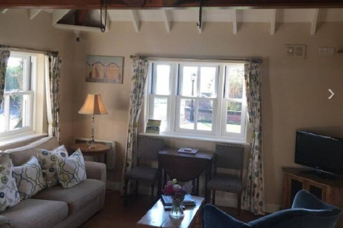 A cozy living room with a sofa, a coffee table, and a TV. There are two windows with curtains, a lamp, and a painting on the wall.