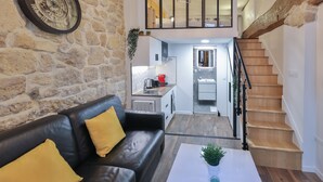 A modern living room with a stone wall, a black leather sofa with yellow cushions, a white coffee table with a plant, and a staircase leading to an upper level.