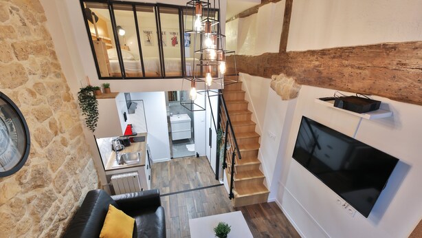 A modern loft apartment with a staircase, a living area with a sofa and coffee table, a kitchen, and a large TV mounted on the wall.