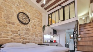 A bedroom with a stone wall, a bed, a small kitchenette, and a staircase leading to an upper level.