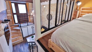 A bedroom with a wooden bed, a window with a view of a stone wall, a staircase, and a small table with a plant.
