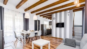 Un salon moderne avec un coin repas, des poutres en bois, un téléviseur à écran plat et une cuisine en arrière-plan.