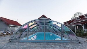 Une piscine intérieure à laquelle on accède par des parois vitrées, surmontée d’un toit incurvé, entourée d’une zone pavée et avec des immeubles d’habitation en arrière-plan.