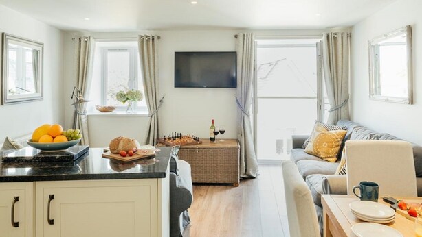A modern kitchen with a dining area, a flat-screen TV, and a large window with curtains. - 8 Sunday School Court (St Ives)