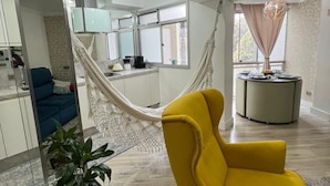 Un salon moderne avec un fauteuil jaune, un hamac, un coin cuisine et une table à manger surmontée d’un lustre.