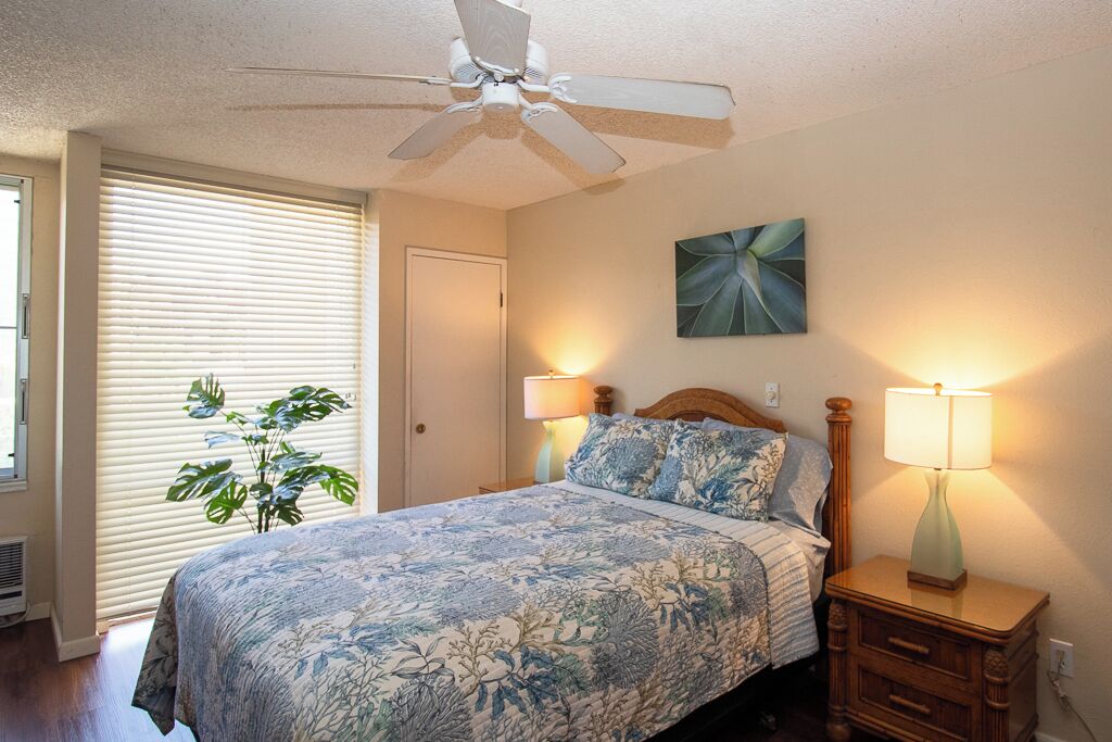 Quarto com cama, duas luminárias de cabeceira, ventilador de teto, janela com persianas e uma planta.