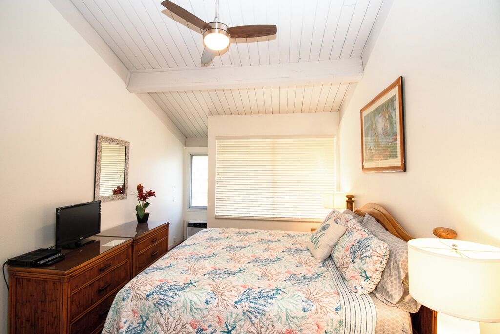 Une chambre à coucher avec un lit, un ventilateur de plafond, un téléviseur posé sur une commode et une fenêtre avec des stores.
