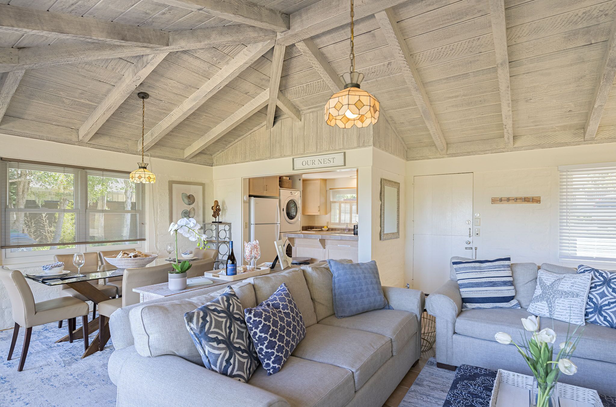 A spacious living area with a high ceiling and exposed beams, a dining table with chairs, a sofa with patterned cushions, and a kitchen area visible through an open door.
