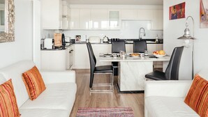Una cocina moderna con armarios blancos, una mesa de comedor y sillas negras. En la sala de estar, hay un sofá blanco con almohadas naranjas.