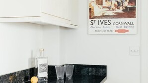 Una cocina con encimera negra, una botella de licor y dos vasos. Un cartel antiguo de St Ives, Cornwall, está en la pared.