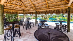 An outdoor seating area with tables and chairs, a thatched roof, and a pool in the background. - Awesome Daydreamer Home, Pool, Waterpark, Slides, Lazy River, near all Parks (Kissimmee)