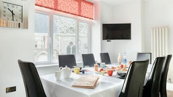 Une salle de conférence avec une table blanche, des chaises noires, une horloge, un téléviseur et une fenêtre donnant sur des immeubles.