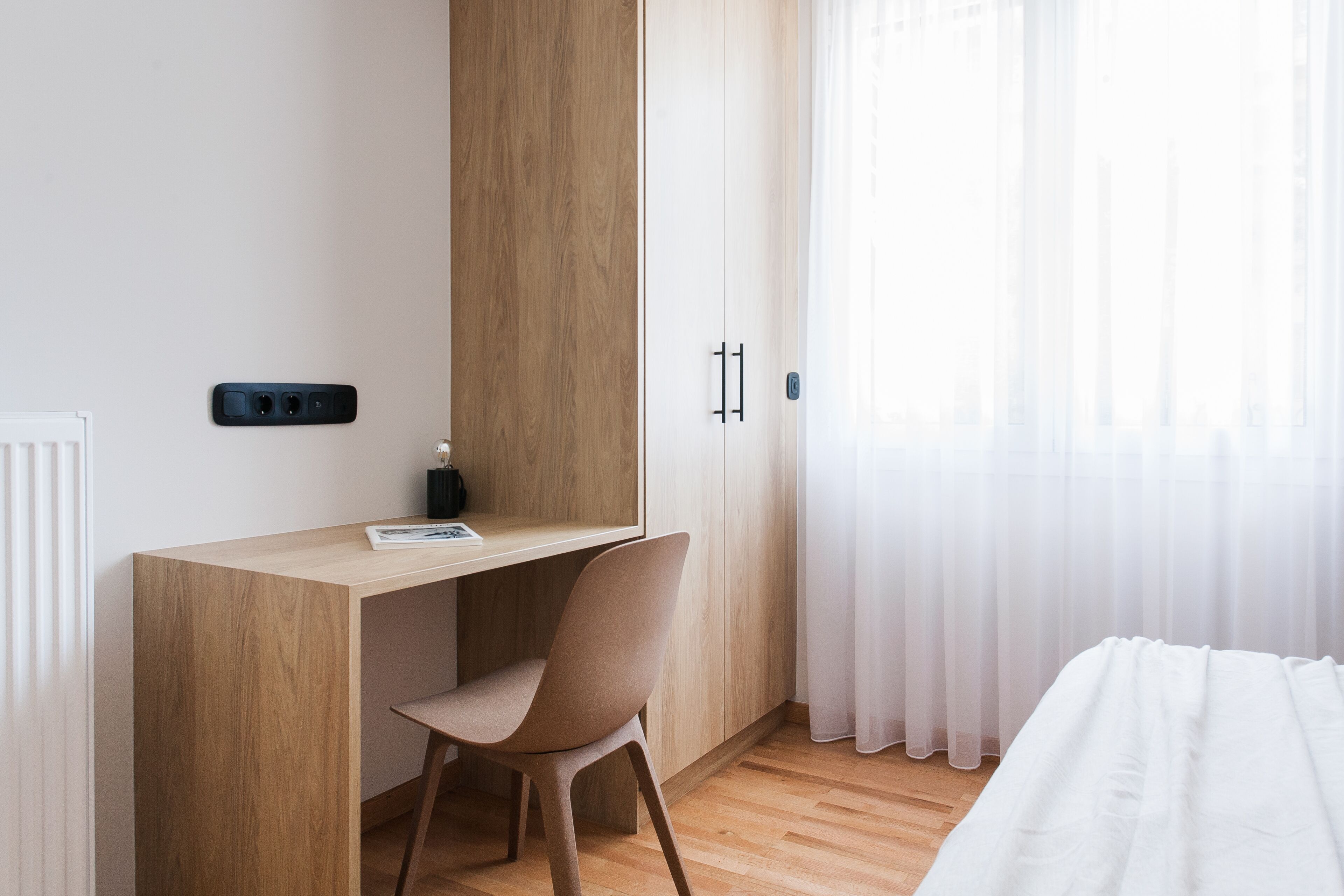 A modern bedroom with a wooden desk, a chair, a bed, and a window with sheer curtains.